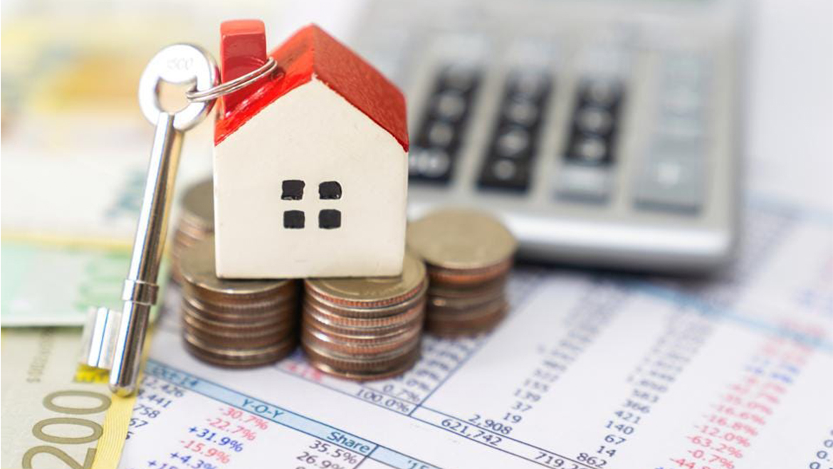 A house figurine with a key placed on piles of change. This is on a table with a calculator behind it and bills under the change.