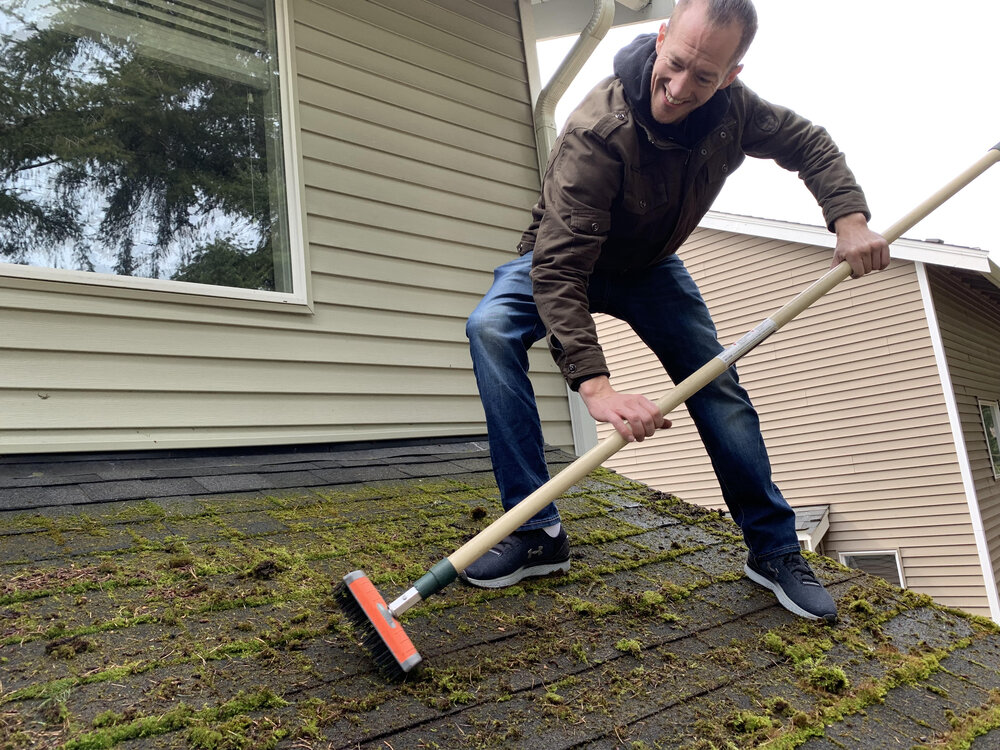 A man standing on the roof using a broom to sweep moss growth