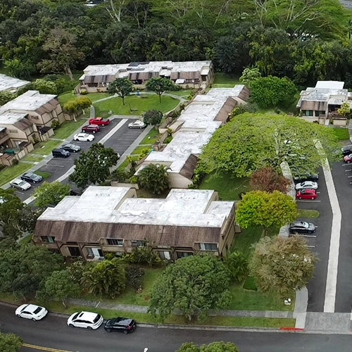There are a series of commercial buildings with several parking lots with cars parked in them. These are surrounded by a variety of trees.